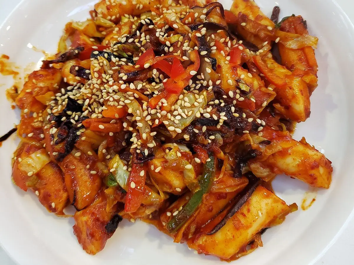 Plato blanco de tteokbokki en salsa roja picante con col, cebolleta y semillas de sésamo