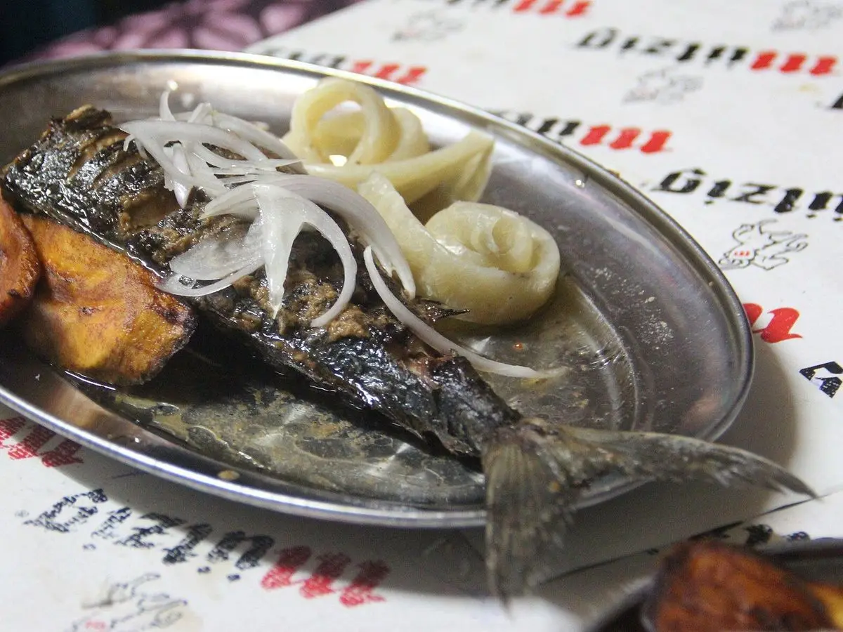 Pescado braseado con cebolla, plátano frito y barritas de yuca en una bandeja metálica