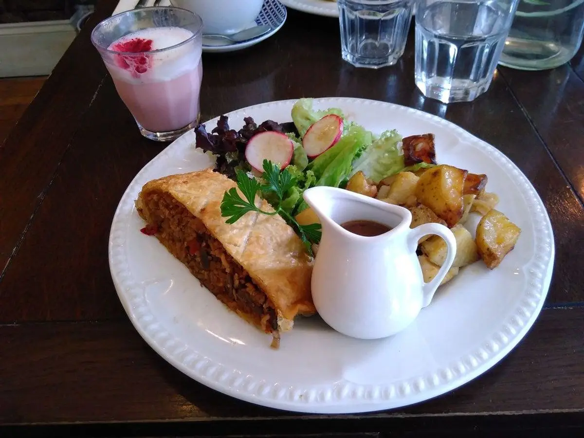 Porción de wellington vegetal en hojaldre con ensalada, patatas asadas y una salsera en un plato blanco