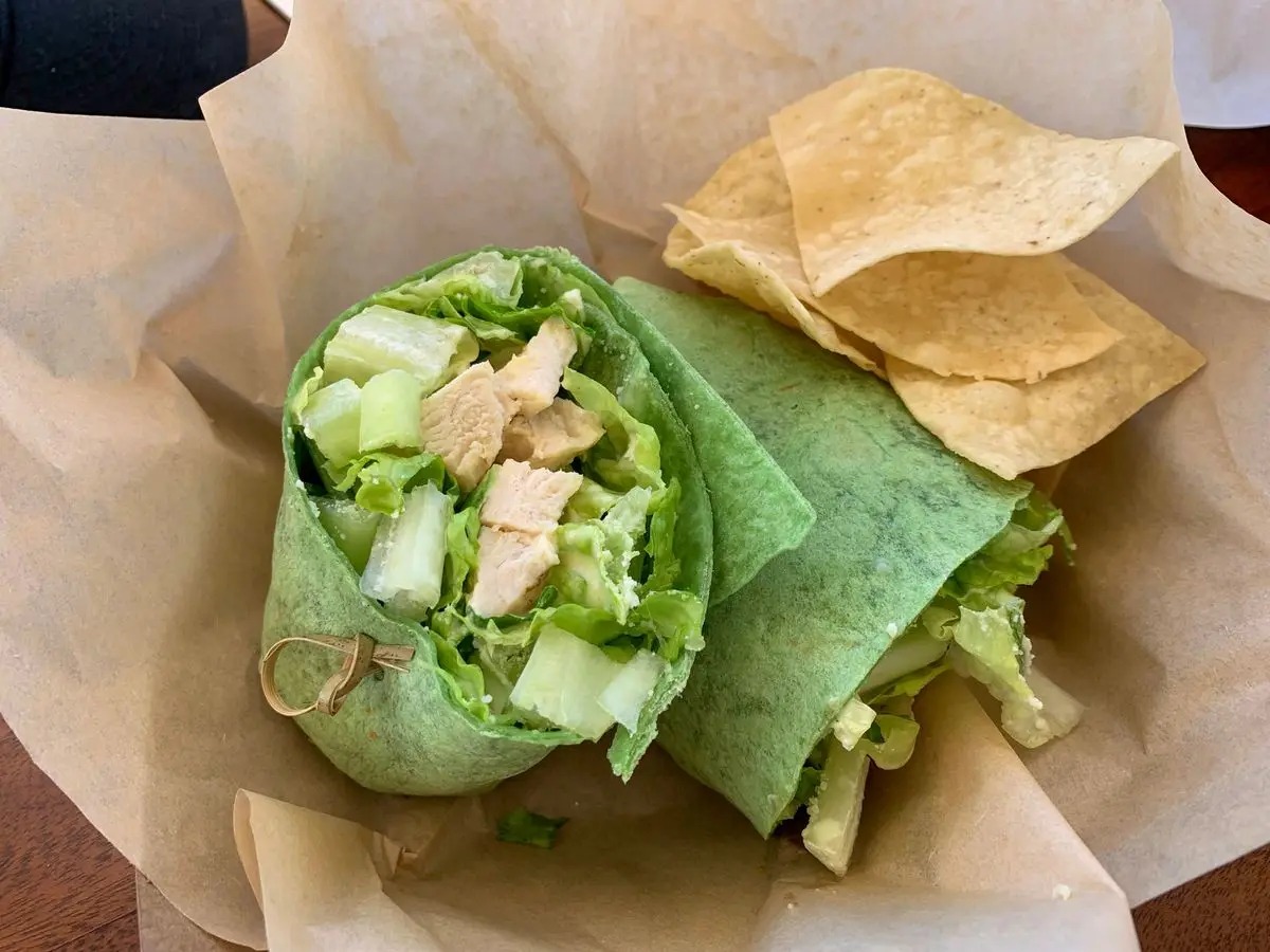 Wrap verde de espinaca cortado por la mitad, relleno de pollo y lechuga, con totopos sobre papel