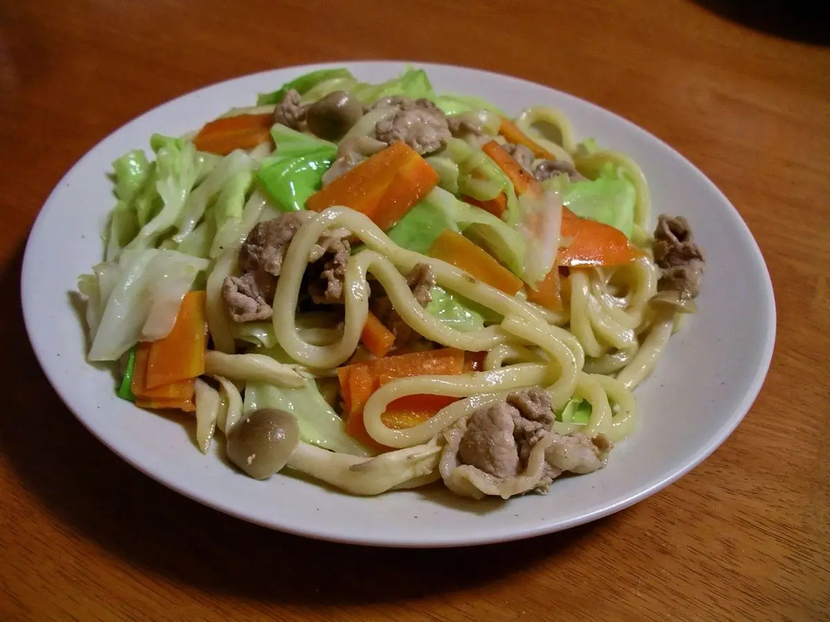Plato de fideos udon salteados con col, zanahoria y tiras de carne sobre una mesa de madera