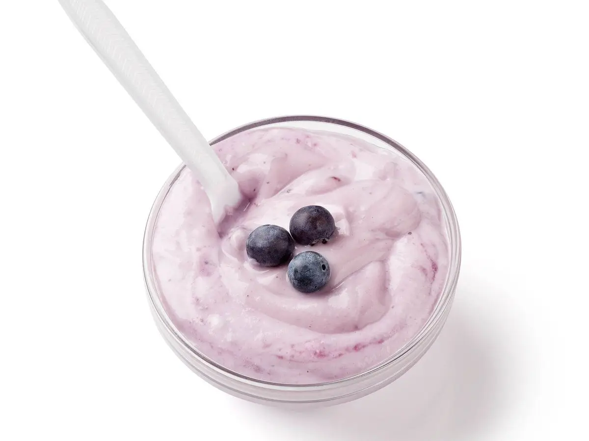 Cuenco transparente de yogur cremoso con tres arándanos por encima y una cuchara