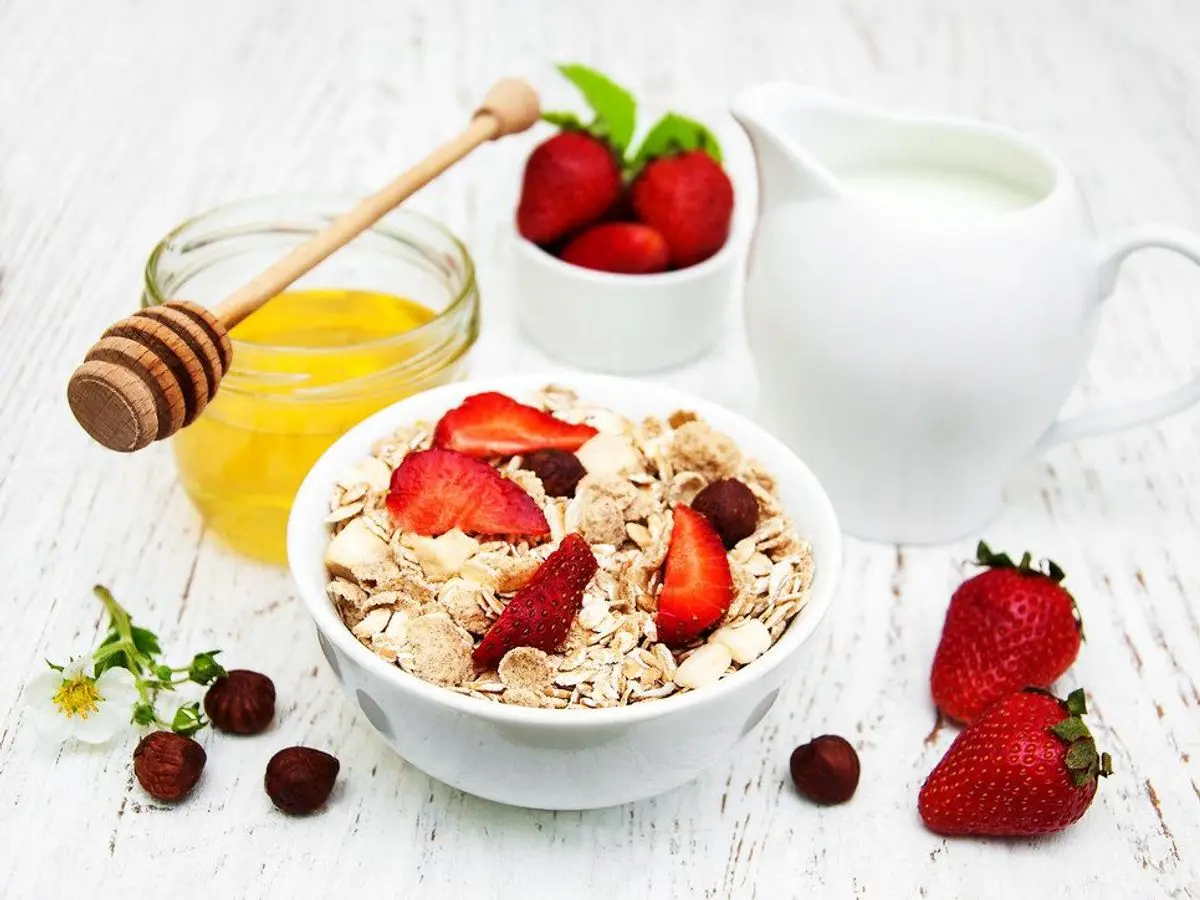 Cuenco de cereales de avena con fresas, junto a un tarro de miel y una jarra de leche